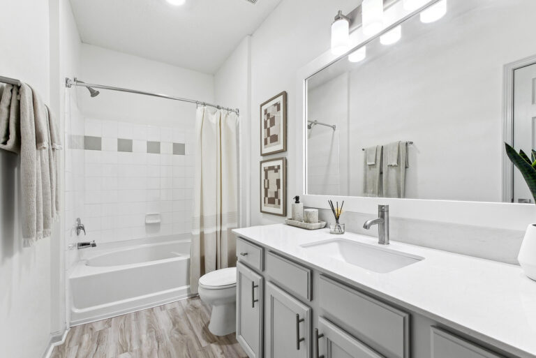 Staged bathroom with white quartz counter tops, gray shaker cabinets, large vanity mirror, toilet, hard-wood style flooring and garden style tub with shower.