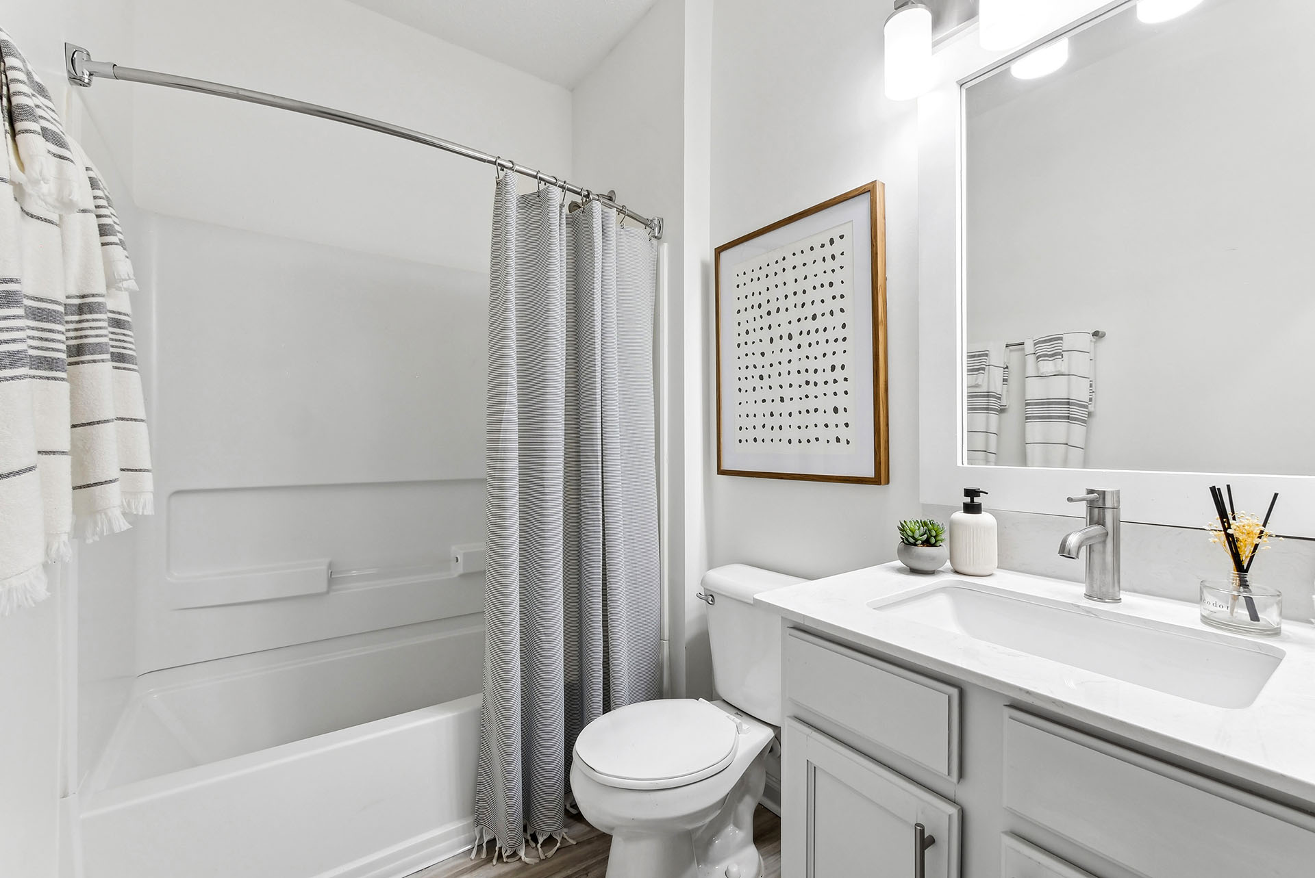 Staged bathroom with white quartz counter tops, gray shaker cabinets, large vanity mirror, toilet, hard-wood style flooring and garden style tub with shower.