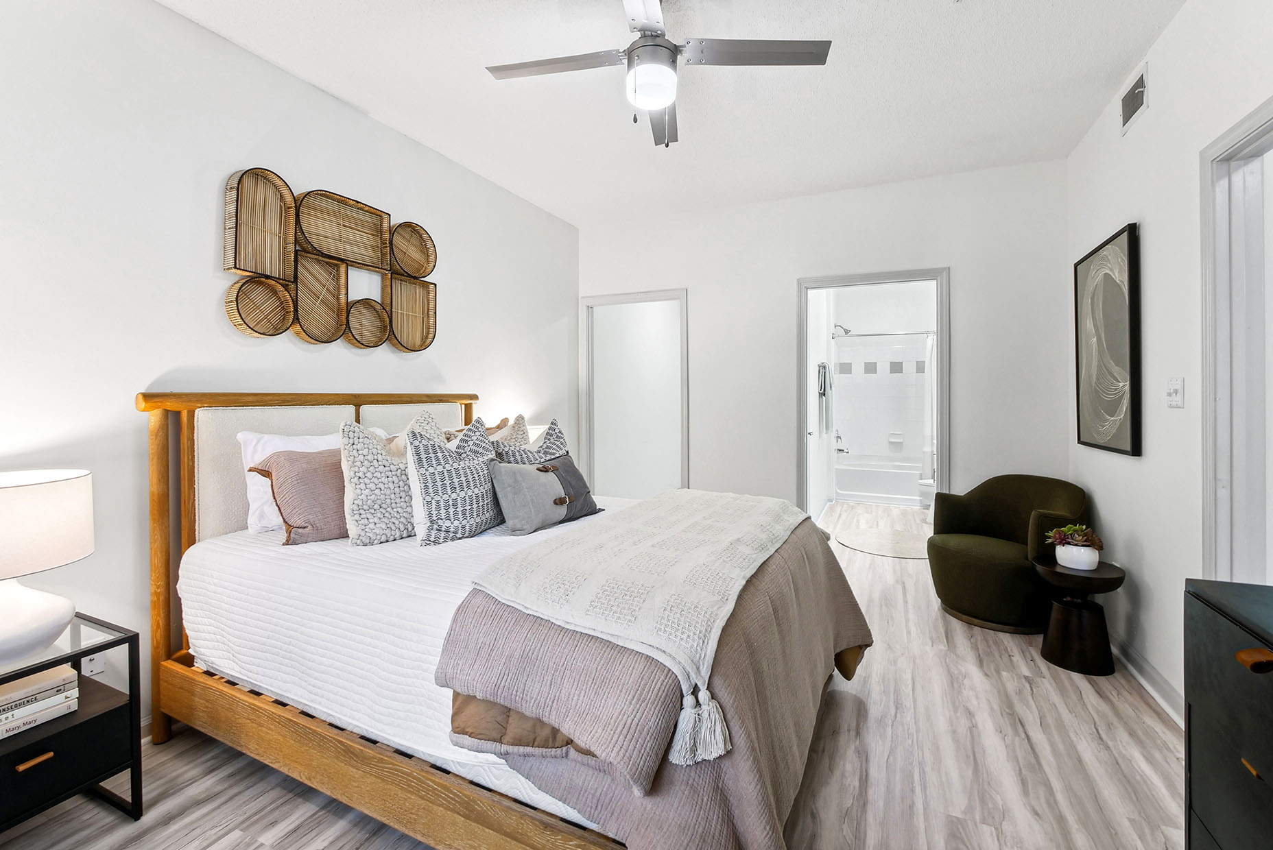 Staged bedroom with bed, hardwood-style flooring, ceiling fan, lounge chair, night stand and ensuite in the background.