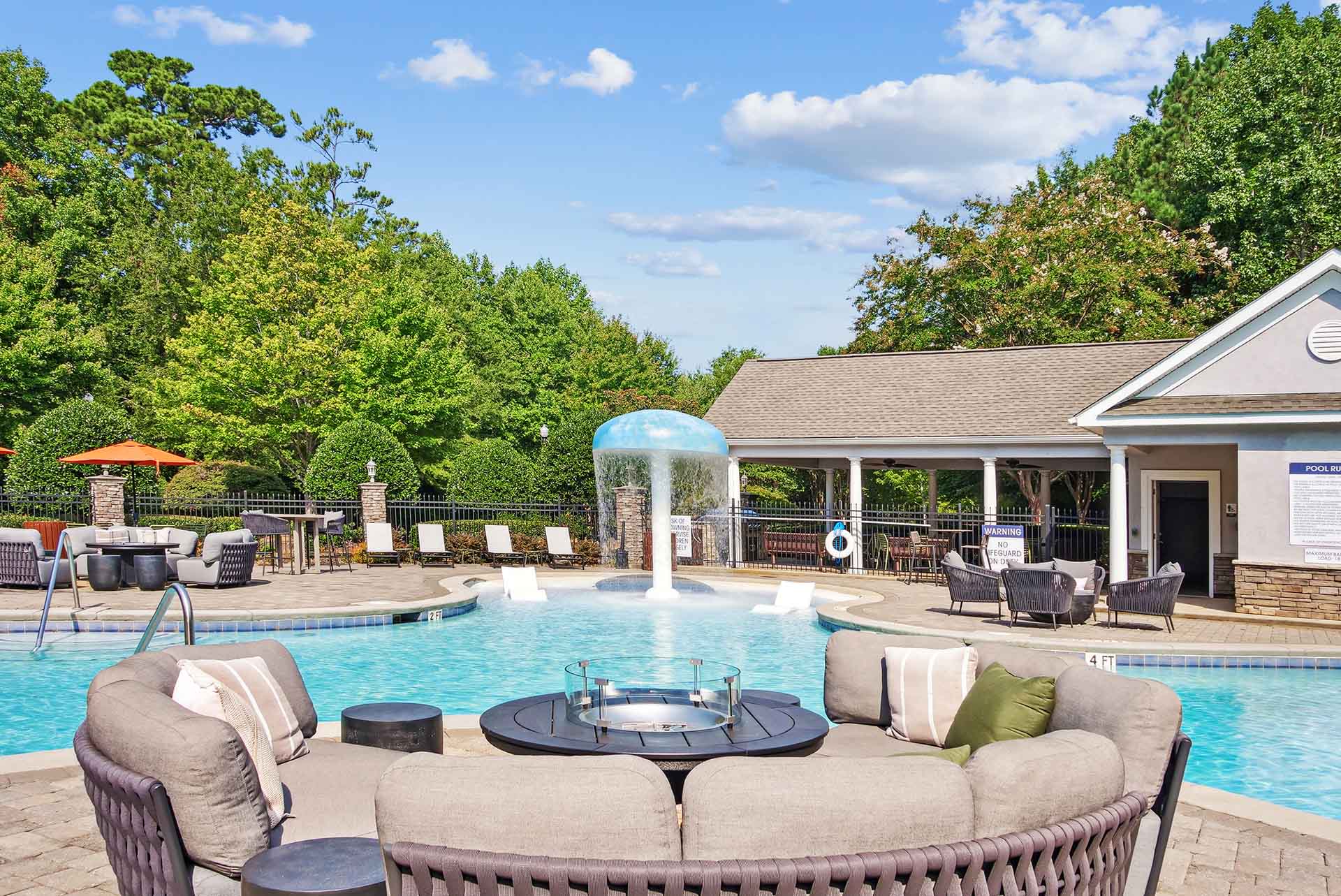Wide view of outdoor fire pit with swimming pool containing a splash zone, large tanning deck and lounge chairs in the backgound surrounded by native landscaping.